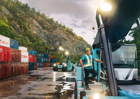 A person standing on the stairs of a lifttruck with nature, containers and another lifttruck in the background.