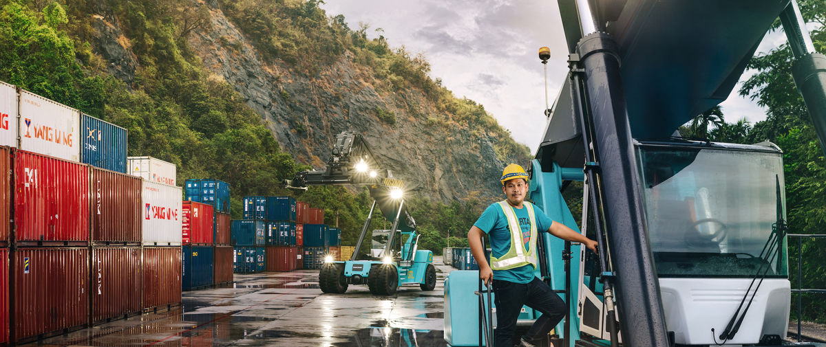 A person standing on the stairs of a lifttruck with nature, containers and another lifttruck in the background.